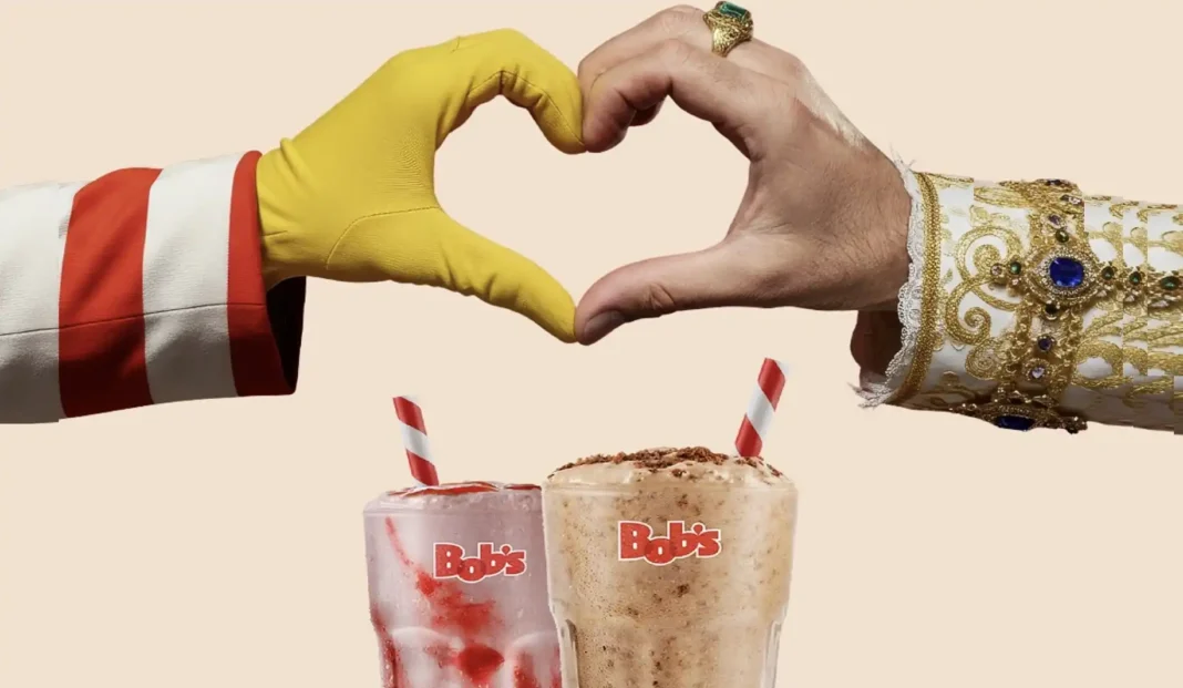 Duas mãos fazendo um formato de coração sobre dois milk-shakes Bob's, um de morango e um de Ovomaltine. Uma das mãos veste uma luva amarela e uma manga listrada em vermelho e branco. A outra mão, com anel e pulseira, veste uma manga branca bordada com dourado e gemas azuis.