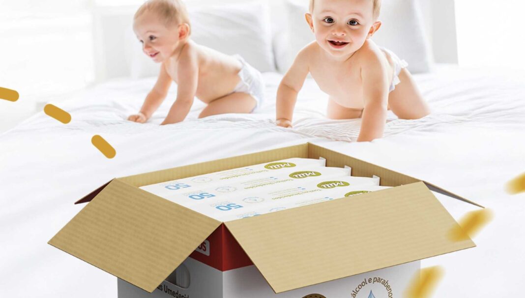 Dois bebês sorridentes engatinham em uma cama branca, com uma caixa de produtos Mili Fralda em primeiro plano.