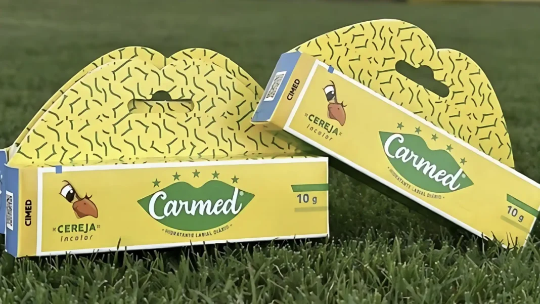 Duas caixas de Carmed Cereja, com embalagem amarela e design de lábios estilizados, sobre a grama.