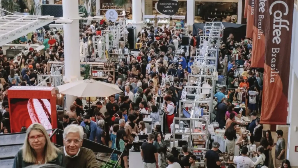 Uma grande convenção de café em um espaço interno lotado de pessoas, com várias barracas e estandes. No primeiro plano, duas pessoas mais velhas sorriem para a câmera. Um grande banner marrom com "Coffee Brasília" em texto vertical pode ser visto à direita.