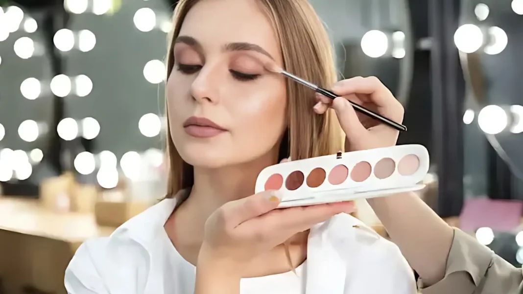A imagem mostra uma sessão de maquiagem, onde uma mulher (presumivelmente a maquiadora) está aplicando sombra nos olhos de outra mulher. A paleta de sombras em destaque parece ser a 'Marc Jacobs Beauty Eye-conic Longwear Eyeshadow Palette Fantascene 790', com seus tons terrosos e rosados. A cena transmite a ideia de um processo de embelezamento profissional, possivelmente em um ambiente de salão de beleza ou estúdio.
