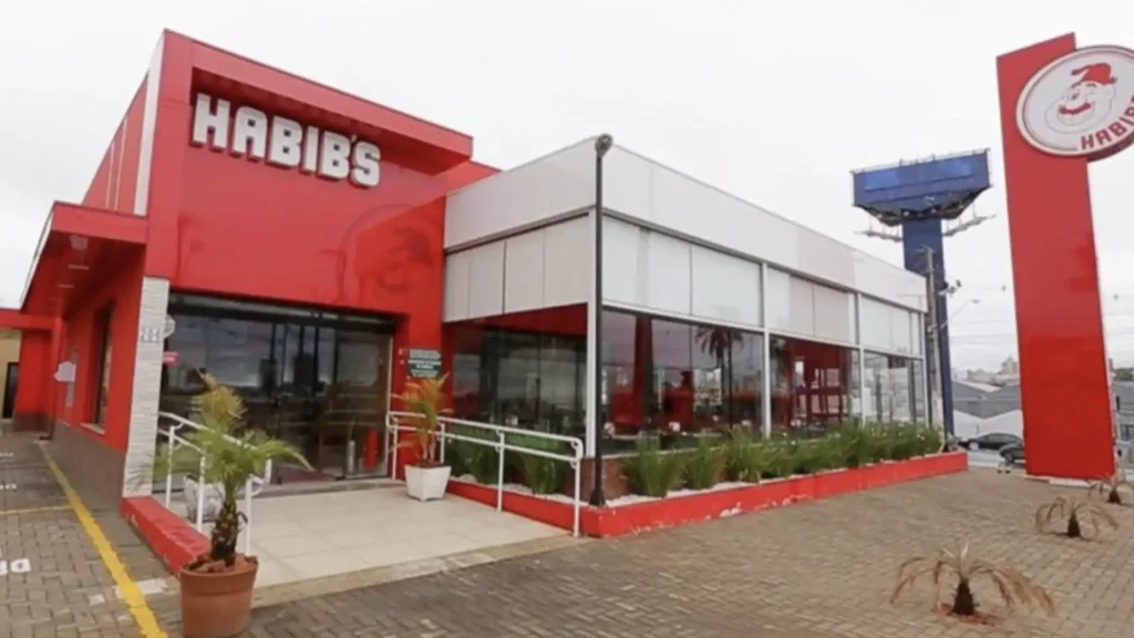 Uma foto externa de um restaurante Habib's, com a fachada em vermelho vibrante e grandes janelas de vidro. A frente do restaurante possui um letreiro grande com o nome "HABIB'S" em branco. À direita da imagem, há uma placa alta com o logotipo do Habib's, que é um Papai Noel estilizado. Há algumas plantas pequenas em vasos na calçada em frente ao restaurante. O céu parece nublado.