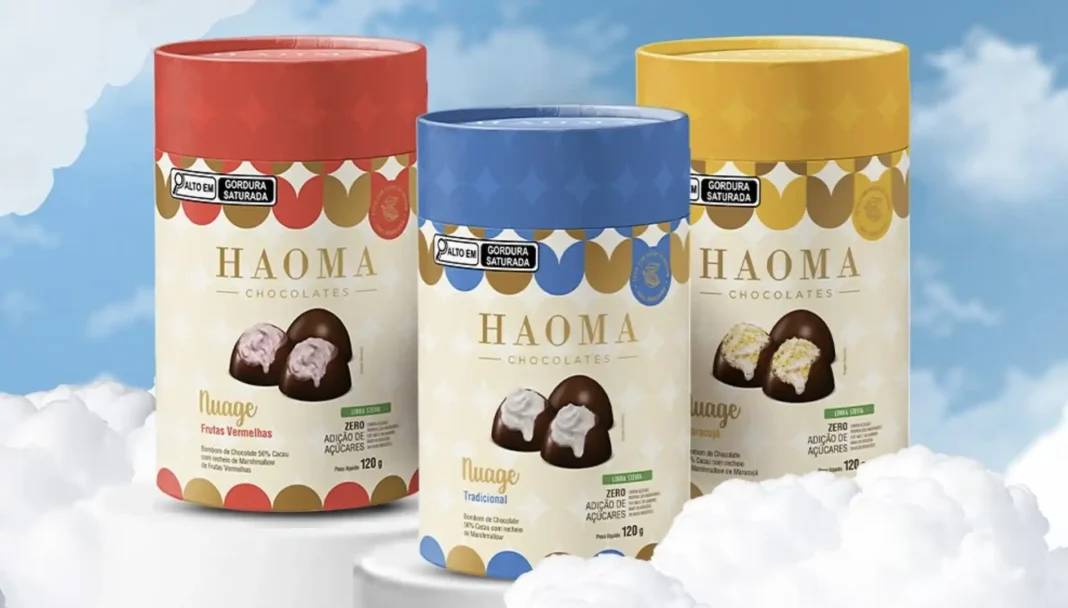 Três embalagens cilíndricas de chocolates gourmet da linha 'Nuage' da Haoma Chocolates. Cada embalagem possui um rótulo com a marca 'HAOMA CHOCOLATES' e o nome da linha 'Nuage', com imagens dos bombons e informações sobre o sabor. À esquerda, uma embalagem vermelha para o sabor 'Frutas Vermelhas' com bombons de chocolate com recheio rosa. No centro, uma embalagem azul para o sabor 'Tradicional' com bombons de chocolate com recheio branco. À direita, uma embalagem amarela para o sabor 'Maracujá' com bombons de chocolate com recheio amarelo. Todas as embalagens exibem selos de 'ALTO EM GORDURA SATURADA' e 'ZERO ADIÇÃO DE AÇÚCARES'. O fundo da imagem simula um céu azul com nuvens brancas, sugerindo leveza e suavidade.