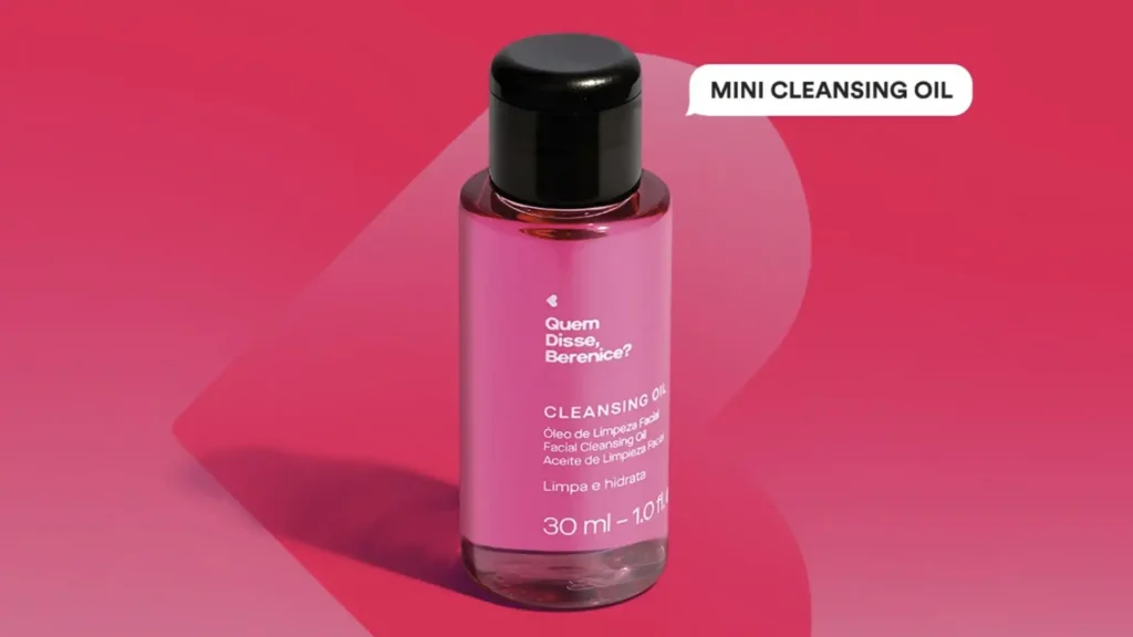 Uma pequena garrafa rosa de Óleo de Limpeza Facial da Quem Disse, Berenice? com uma tampa preta, em um fundo rosa. A garrafa tem um rótulo que diz "CLEANSING OIL" e "30 ml - 1.0 fl.oz". O texto "MINI CLEANSING OIL" está em destaque no canto superior direito da imagem.