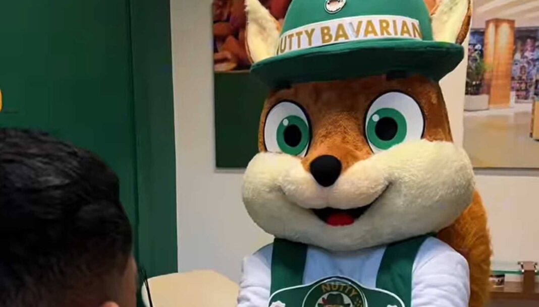 Mascote da Nutty Bavarian, um simpático esquilo de avental e chapéu bávaro, sorrindo enquanto segura um cone de nuts caramelizados.