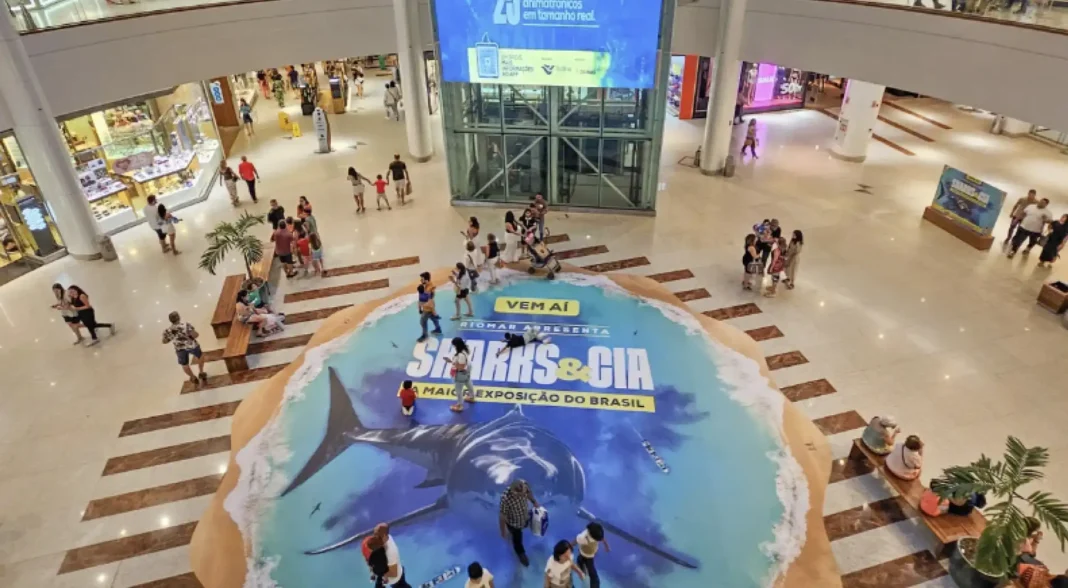 Uma vista de cima de um shopping center movimentado, com um grande display redondo no centro do piso térreo. O display tem um tema de praia, com areia e água azul-clara, e apresenta uma imagem de um grande tubarão. O texto no display diz 'VEM AÍ RIOMAR APRESENTA SHARKS & CIA A MAIOR EXPOSIÇÃO DO BRASIL'. Pessoas estão andando ao redor do display e nas lojas ao fundo.