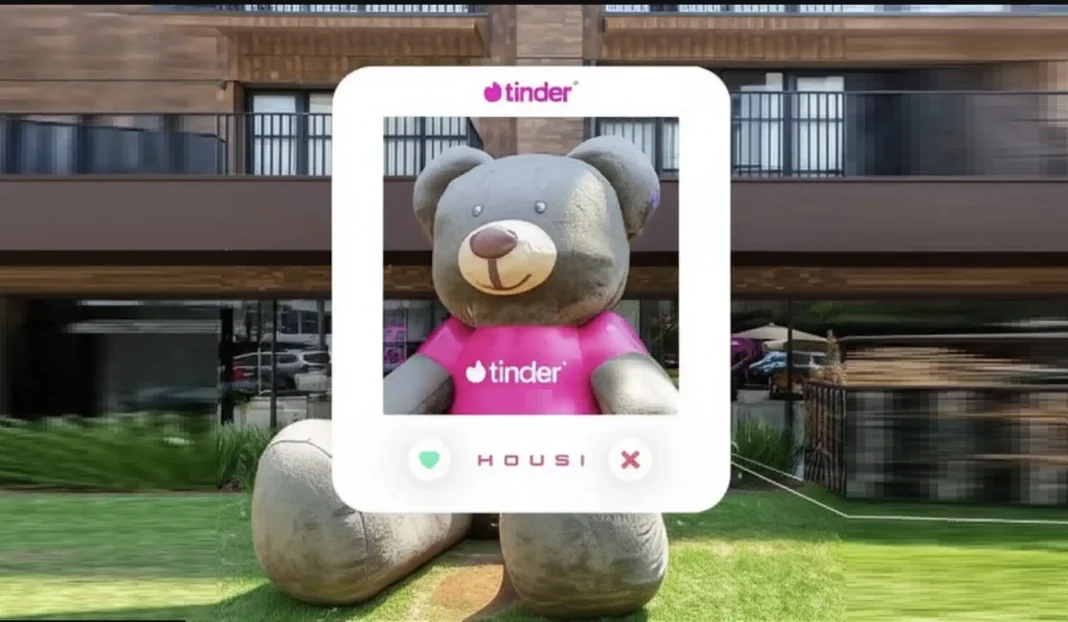 Um ursinho de pelúcia gigante, com a camiseta rosa do Tinder, está sentado na grama em frente a um prédio moderno, simulando um perfil do Tinder com os logos do Tinder e Housi.
