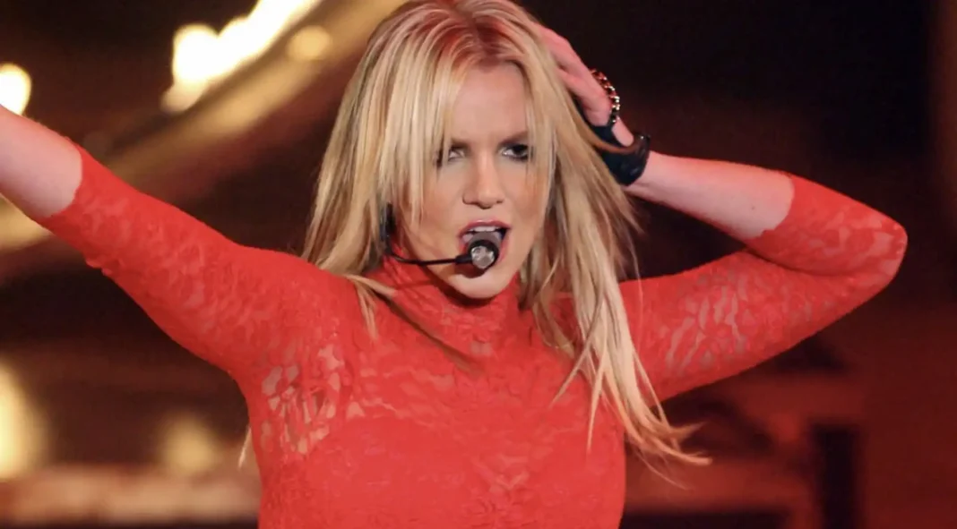 Britney Spears se apresenta no palco, com as mãos levantadas, vestindo uma blusa de renda vermelha.