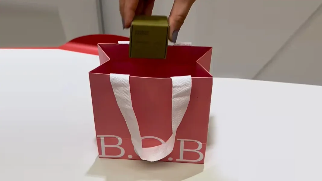 Uma mão colocando uma caixa verde dentro de uma sacola de presente rosa com as letras 