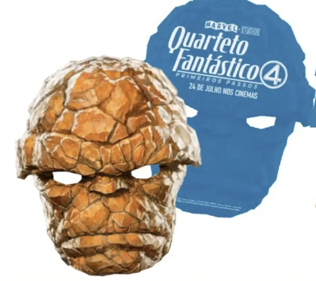 
Uma máscara de brinquedo do Coisa (The Thing) do Quarteto Fantástico em primeiro plano, feita de um material texturizado que se assemelha a pedras alaranjadas e irregulares. A máscara tem furos para os olhos e uma expressão carrancuda. Ao fundo, uma máscara azul lisa com o logo "Marvel Studios Quarteto Fantástico" e a frase "Primeiros Passos 24 de Julho Nos Cinemas" em branco.