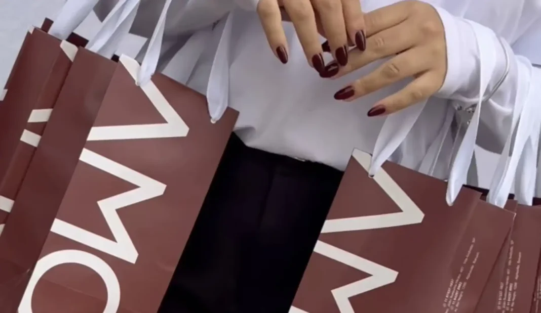 Mãos femininas com unhas bordô segurando sacolas marrons com o logo 'MO'.