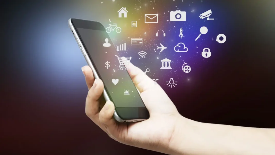 A mão de uma pessoa segura um smartphone, do qual emanam diversos ícones brancos e brilhantes representando várias aplicações e funcionalidades digitais, sobre um fundo escuro com tons roxos e amarelos.