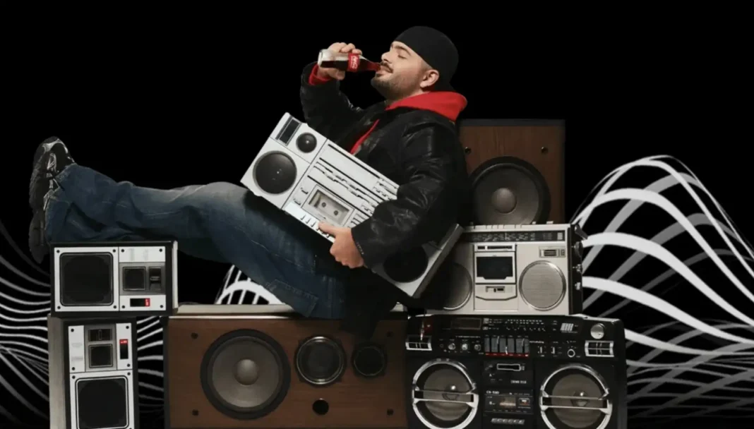 Um homem com um gorro preto, jaqueta de couro e moletom vermelho, deitado de lado sobre pilhas de rádios antigos e boomboxes, segurando um boombox branco e bebendo uma Coca-Cola. Ao fundo, um design abstrato em preto e branco.