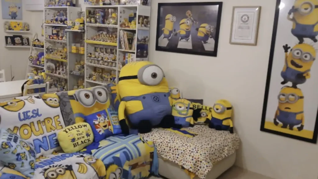 
Uma foto de um quarto ou sala de estar decorado com uma vasta coleção de itens dos Minions. Há prateleiras brancas repletas de bonecos, brinquedos e objetos relacionados aos personagens amarelos. Um sofá está coberto com almofadas e cobertores dos Minions, incluindo uma almofada amarela com a frase "YELLOW IS THE NEW BLACK". Uma cama ou divã também possui lençóis com estampas dos Minions e vários bonecos de pelúcia dos Minions de tamanhos variados, incluindo um grande no centro. Na parede, há quadros com pôsteres dos Minions, um deles em estilo de capa de álbum, e um certificado do Guinness World Records. A iluminação é clara e o ambiente é predominantemente nas cores amarelo, azul e branco, em referência aos personagens.