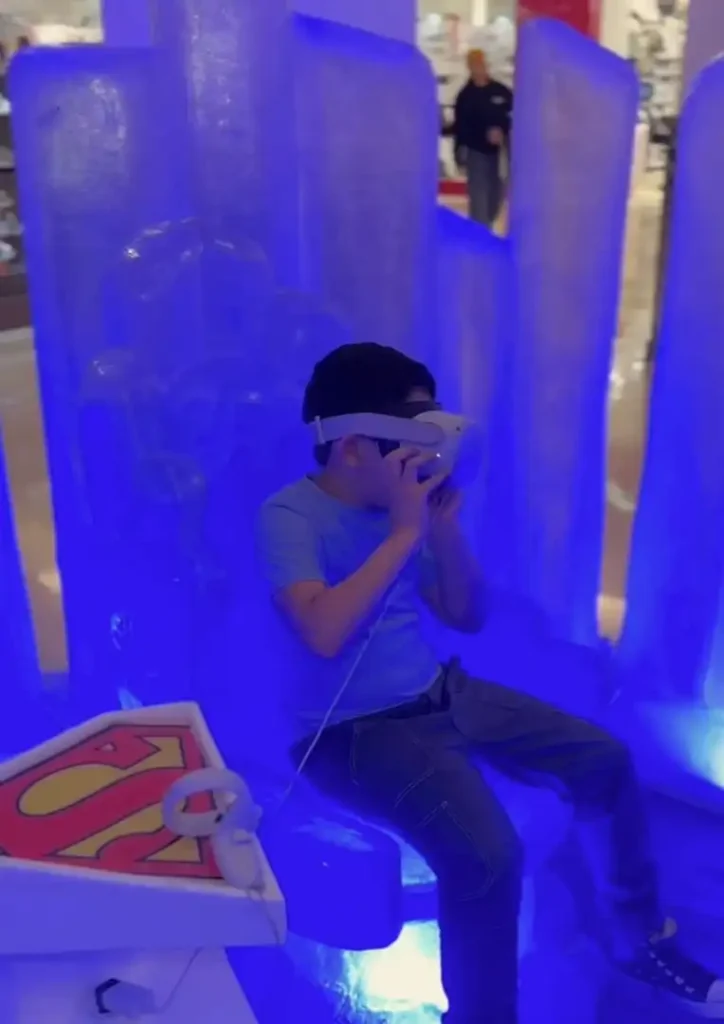 
Um garoto de camiseta azul e calça jeans escura está sentado em um trono que parece ser feito de gelo azul iluminado. Ele está usando um óculos de realidade virtual branco e segurando controles brancos nas mãos. À sua esquerda, há um pedestal com o logotipo vermelho e amarelo do Superman. O ambiente parece ser um espaço interno, possivelmente um shopping, com uma pessoa caminhando ao fundo desfocado.