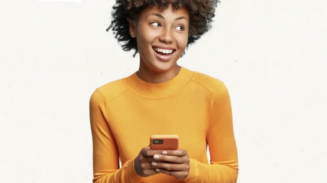 Mulher jovem e feliz, de pele morena e cabelo crespo, usando uma blusa laranja e segurando um smartphone coral.