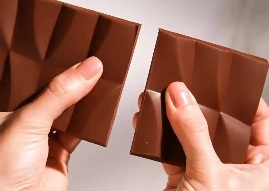 
Uma barra de chocolate marrom está sendo partida ao meio por duas mãos com as unhas pintadas de rosa.