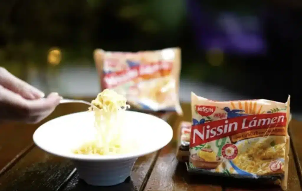 Uma mão segura um garfo com macarrão instantâneo em um prato branco. Ao lado do prato, há um pacote de Nissin Lámen com o sabor "Cuzcuz com Calabresa". Outros pacotes de macarrão estão desfocados ao fundo.