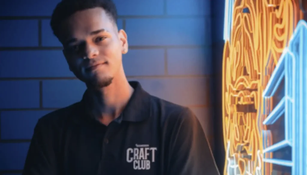 Um homem jovem de pele clara com cabelo curto e escuro, vestindo uma camisa polo preta com o logo "Craft Club", olha para a câmera com um sorriso leve. O fundo apresenta uma parede de tijolos azuis parcialmente iluminada e uma luz de neon amarela e azul desfocada à direita.