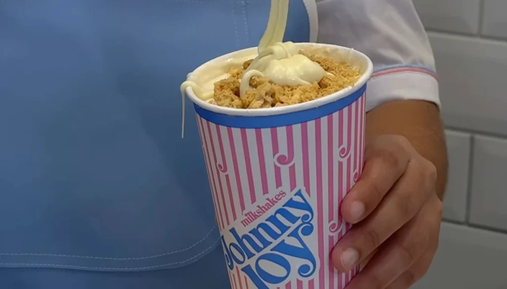 Close-up de um milkshake Galaktico da Johnny Joy sendo preparado. Um líquido branco cremoso está sendo despejado sobre o milkshake já preenchido com pedaços de biscoito e uma camada de chantilly. O milkshake está em um copo listrado verticalmente em branco e rosa com detalhes azuis e o logo da Johnny Joy inclinado. Uma mão segura firmemente o copo. Ao fundo, uma pessoa vestindo um uniforme azul claro e branco está parcialmente visível, indicando o preparo do milkshake em um ambiente de serviço de alimentação.