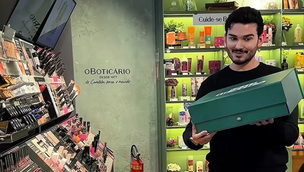 Um homem sorridente segura uma caixa verde em uma loja d'O Boticário, com displays de maquiagem e produtos de beleza ao fundo.