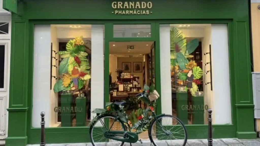 
Uma foto de uma loja verde escura da Granado, com a fachada em madeira e vidro. Há plantas tropicais grandes e coloridas nas vitrines de cada lado da porta. Em frente à porta, há uma bicicleta verde escura com uma cesta e flores. O nome "Granado Pharmácias" está escrito em letras douradas acima da porta.