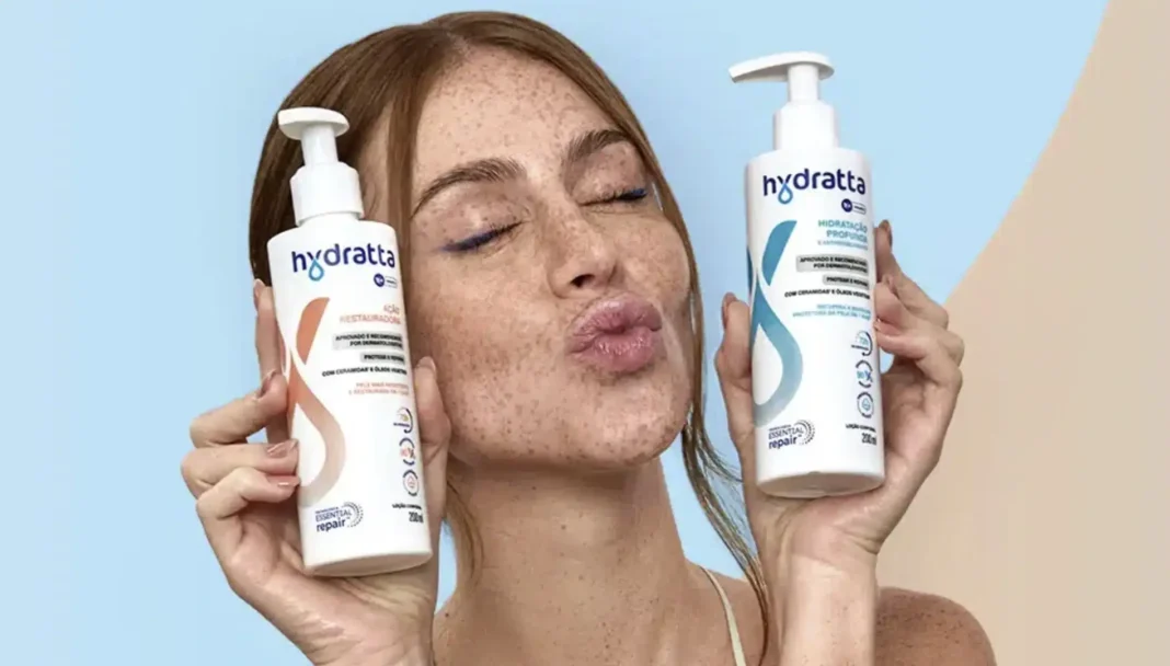 Mari Maria, uma mulher ruiva com sardas e maquiagem nos olhos, segura dois frascos brancos da marca Hydratta.