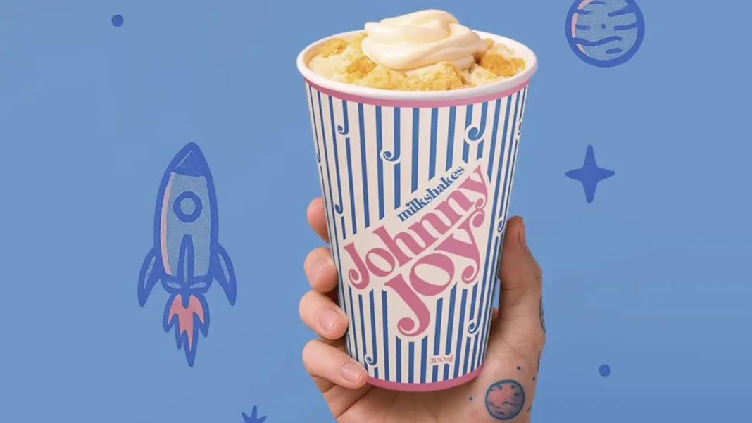 Um milkshake de baunilha cremoso com pedaços de biscoito e cobertura de chantilly, em um copo com listras azuis e brancas da Johnny Joy, sendo segurado por uma mão com tatuagens de planetas. O fundo é azul com desenhos de foguete e estrelas, sugerindo um tema espacial.