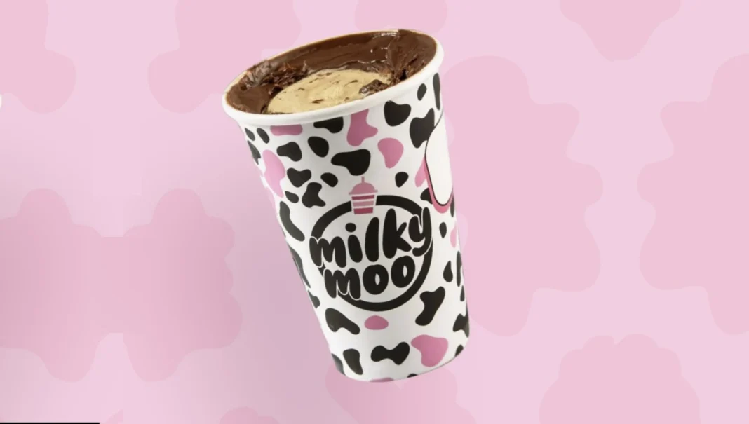 Um copo de milkshake da Milky Moo com estampa de vaca, cheio de milkshake cremoso com cobertura de chocolate e um pedaço de biscoito ou doce por cima, em um fundo rosa claro com bolinhas.