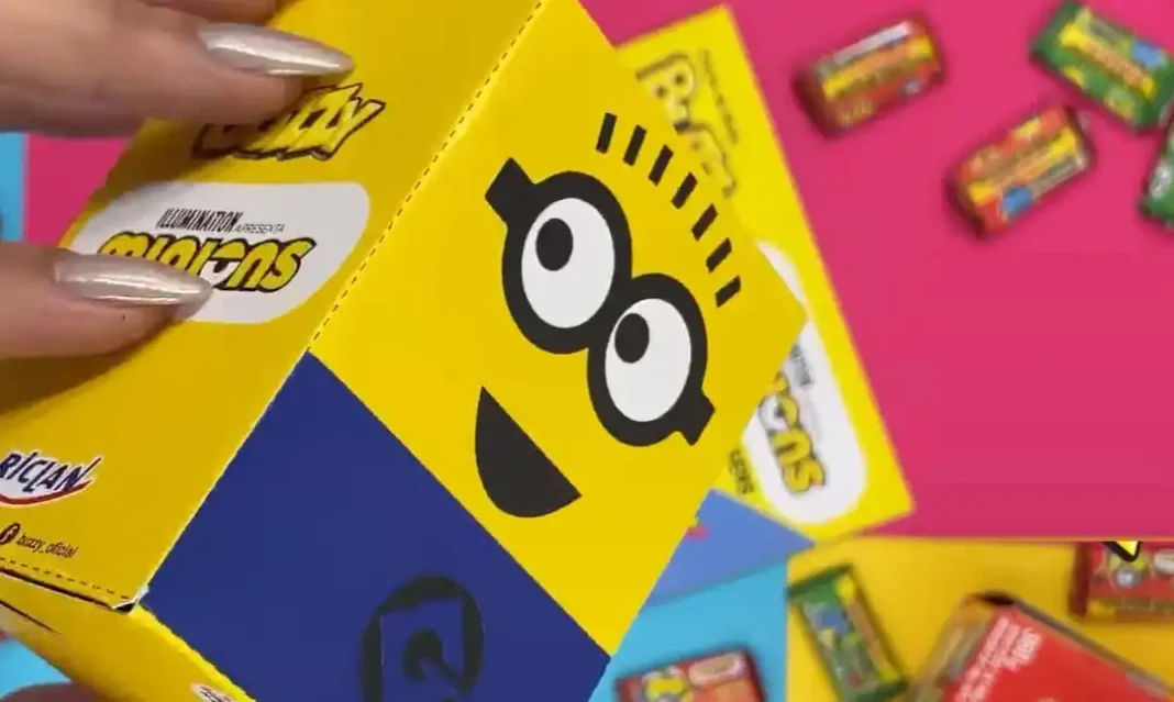 mão segurando uma caixa amarela com tema dos Minions, que parece ser um porta-objetos. A caixa tem o rosto de um Minion feliz, com óculos pretos grandes e um sorriso. A parte inferior da caixa é azul.