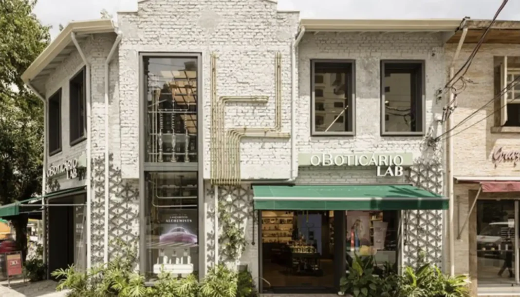 Fachada de tijolos brancos de uma loja da O Boticário Lab com uma grande vitrine e toldo verde, cercada por plantas.