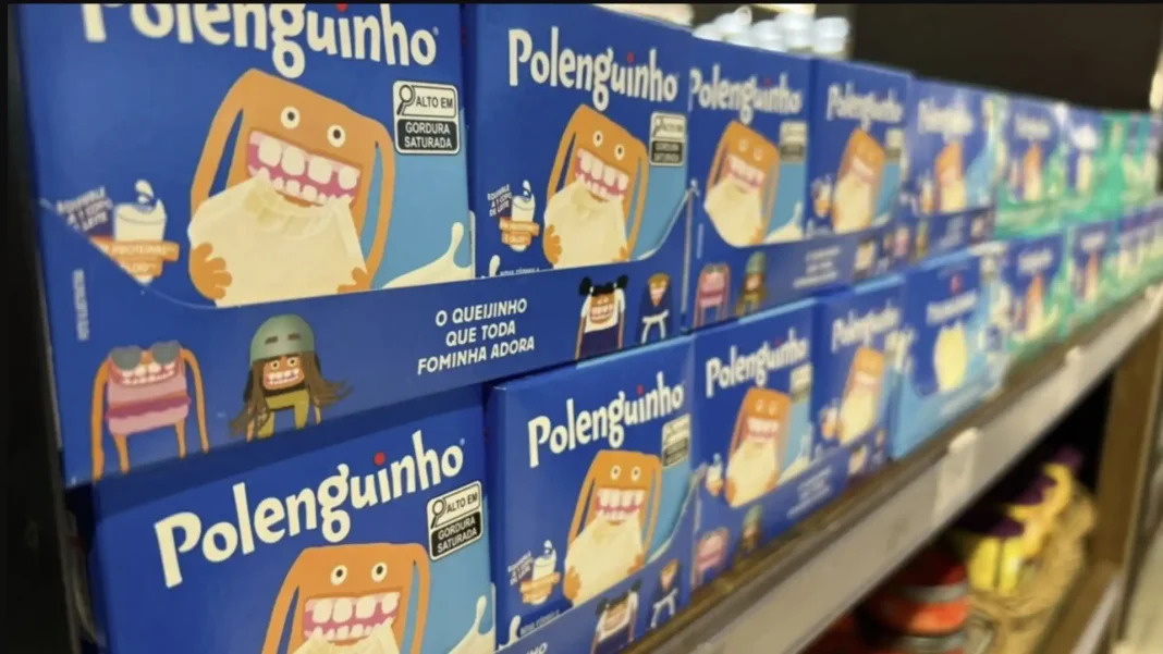 Close-up de múltiplas caixas azuis de Polenguinho empilhadas em uma prateleira de supermercado. O design da embalagem apresenta um monstrinho laranja sorridente com grandes dentes, segurando um pedaço de queijo branco cremoso e escorrendo leite. O texto principal na caixa diz 'Polenguinho' e 'O QUEIJINHO QUE TODA FOMINHA ADORA'