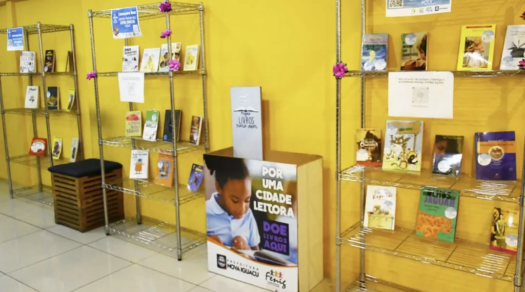 Uma imagem mostra um ambiente com paredes amarelas e estantes de metal cheias de livros. Ao centro, um totem em forma de livro exibe a imagem de uma criança lendo e a frase "POR UMA CIDADE LEITORA DOE LIVROS AQUI".