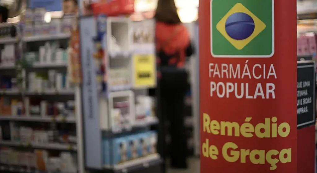 
Um cartaz vertical vermelho em uma farmácia anuncia "Farmácia Popular" e "Remédio de Graça" em letras brancas e amarelas, respectivamente, com uma bandeira brasileira no topo. Ao fundo, uma pessoa desfocada anda por entre as prateleiras de produtos.