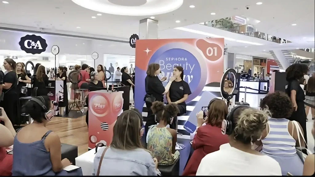 
Uma visão de um evento da Sephora em um shopping, com uma cabine de maquiagem ao centro, onde uma maquiadora aplica maquiagem em uma mulher sentada. Pessoas, principalmente mulheres, estão reunidas ao redor, algumas assistindo e outras participando de atividades relacionadas à beleza. O ambiente é bem iluminado e parece ser um espaço de varejo movimentado, com outras lojas e escadas visíveis ao fundo.