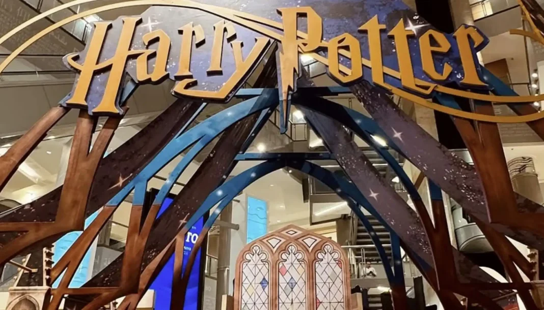 Um banner promocional da exposição de Harry Potter no Shopping Tietê Plaza. A imagem mostra a entrada da exposição, com a logo 'Harry Potter' em destaque no topo, sobre uma estrutura de madeira escura e detalhes em azul, lembrando um castelo ou uma catedral gótica, com algumas estrelas e pontos de luz.