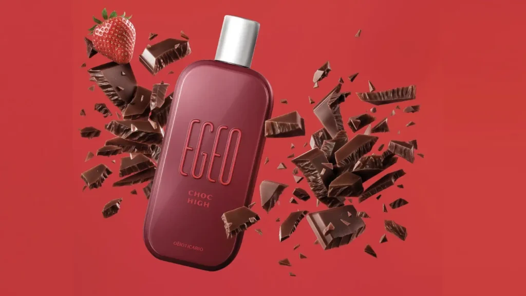 
Uma garrafa vermelha escura do perfume "Egeo Choc High" está centralizada contra um fundo vermelho brilhante. Um fluxo de pedaços de chocolate escuro está espalhado ao redor da garrafa, e um morango vermelho brilhante está no canto superior esquerdo.