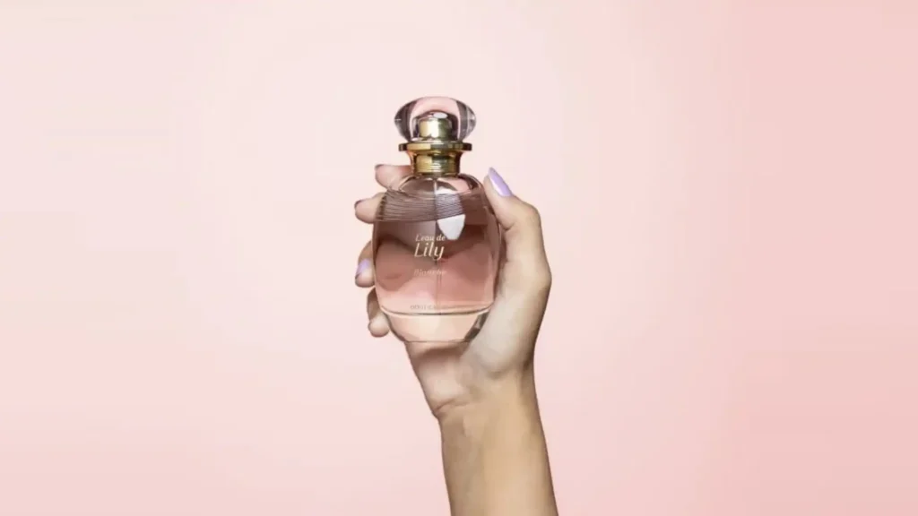 
Uma mão com unhas roxas claras segura uma garrafa de perfume Lily L'eau de Blanche contra um fundo rosa claro. O frasco de perfume tem um corpo transparente que mostra o líquido rosa pálido e uma tampa esférica transparente.