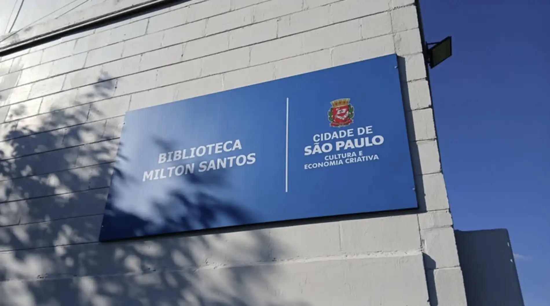 Placa azul retangular fixada em uma parede de tijolos cinza claro, com o texto "BIBLIOTECA MILTON SANTOS" à esquerda e "CIDADE DE SÃO PAULO CULTURA E ECONOMIA CRIATIVA" à direita, separados por uma linha vertical branca. Acima do texto da cidade, há o brasão da cidade de São Paulo em cores.