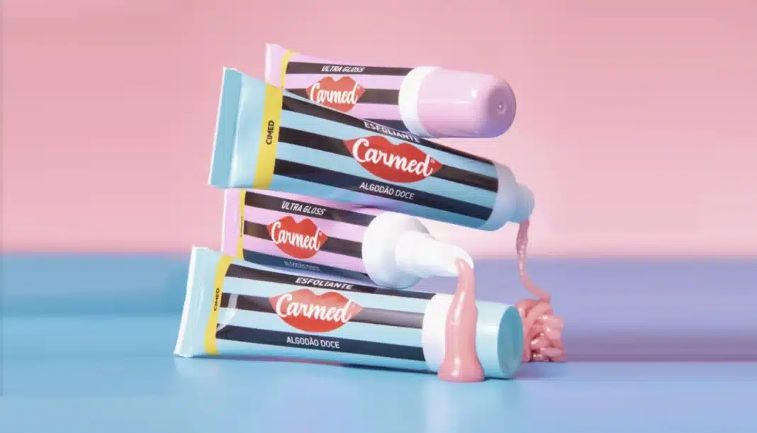 Quatro tubos de produto labial Carmed dispostos em forma de pirâmide, com dois tubos derramando um gel rosa. O fundo é um gradiente de rosa e azul claro.
