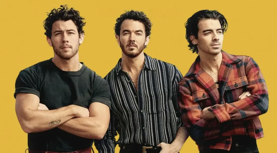 Os Jonas Brothers, da esquerda para a direita, são Nick, Kevin e Joe. Nick está à esquerda, com os braços cruzados, vestindo uma camiseta escura. Kevin está no centro, de frente para a câmera, com uma camisa listrada. Joe está à direita, olhando para o lado, vestindo uma camisa xadrez. O fundo é amarelo sólido.