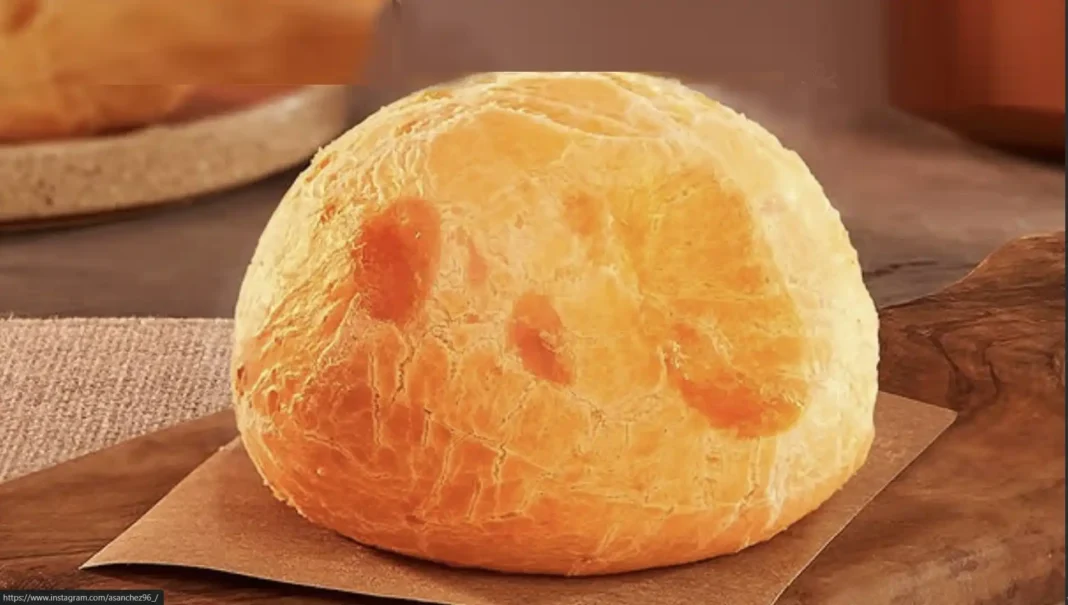 Pão de queijo grande e dourado, servido sobre um pedaço de papel pardo em uma tábua de madeira.