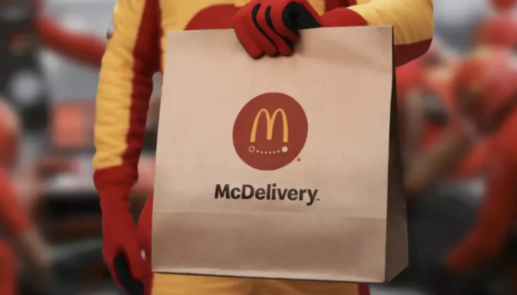 Um close-up de uma pessoa em um traje de piloto de Fórmula 1, vermelho e amarelo, segurando uma sacola de papel da McDelivery com o logotipo do McDonald's.