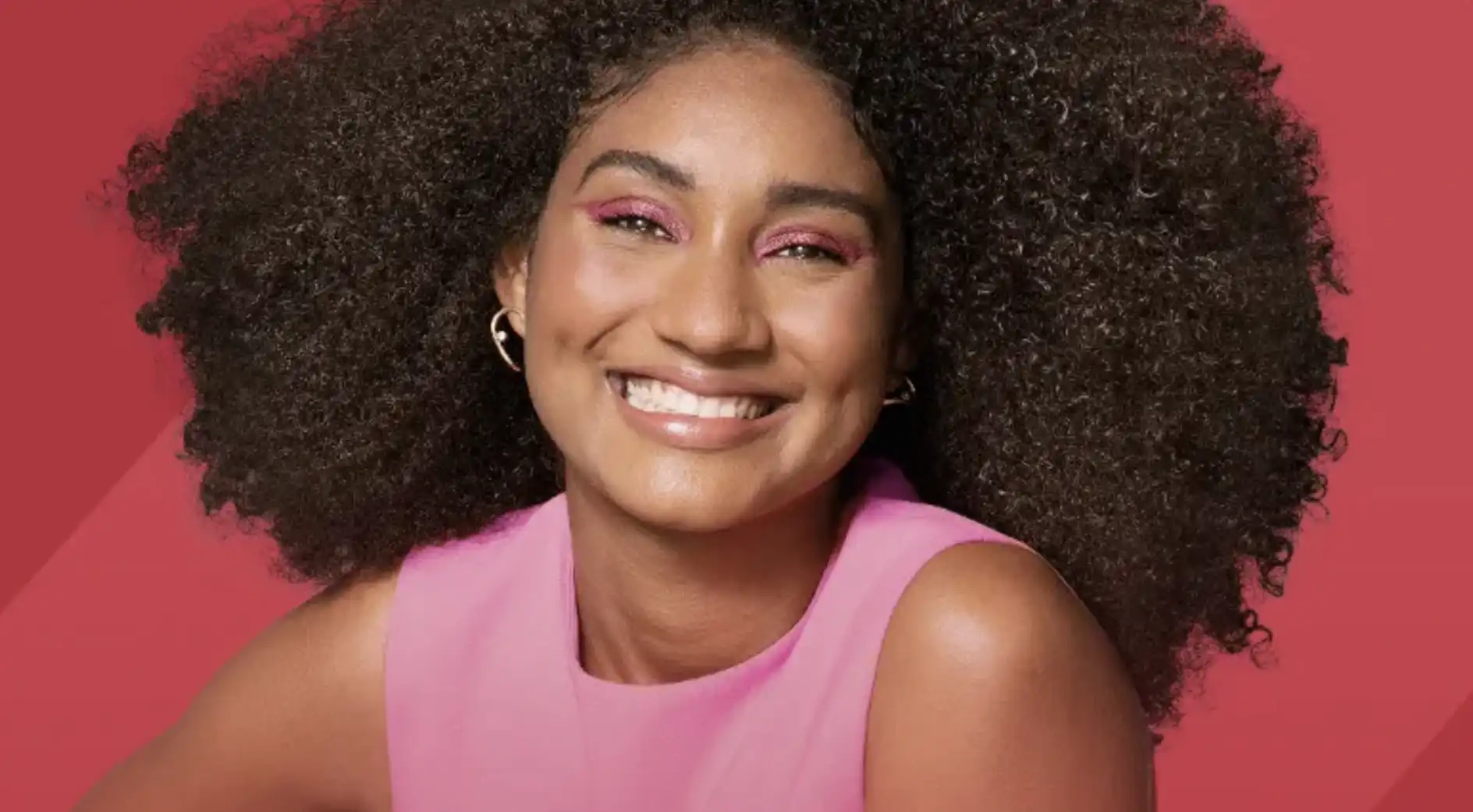 Mulher com cabelo crespo e pele morena sorrindo, usando maquiagem rosa nos olhos e um batom nude brilhante, com fundo vermelho.