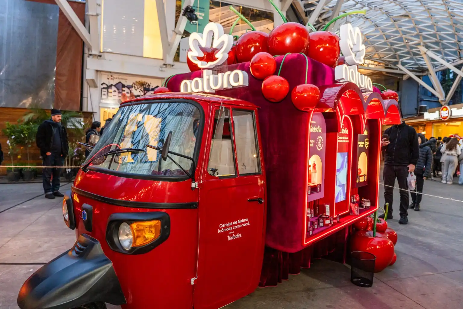 Tuk-tuk vermelho da Natura "Tododia Cereja" em um evento promocional, com cerejas decorativas e vitrines de produtos.