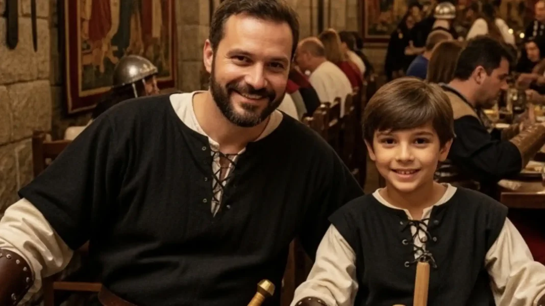 Um homem barbudo sorridente, vestido com uma camisa preta e creme estilo medieval e uma bainha de espada visível em seu cinto, posa ao lado de um menino sorridente, que usa uma roupa medieval semelhante, com uma bainha de espada visível em sua mão.