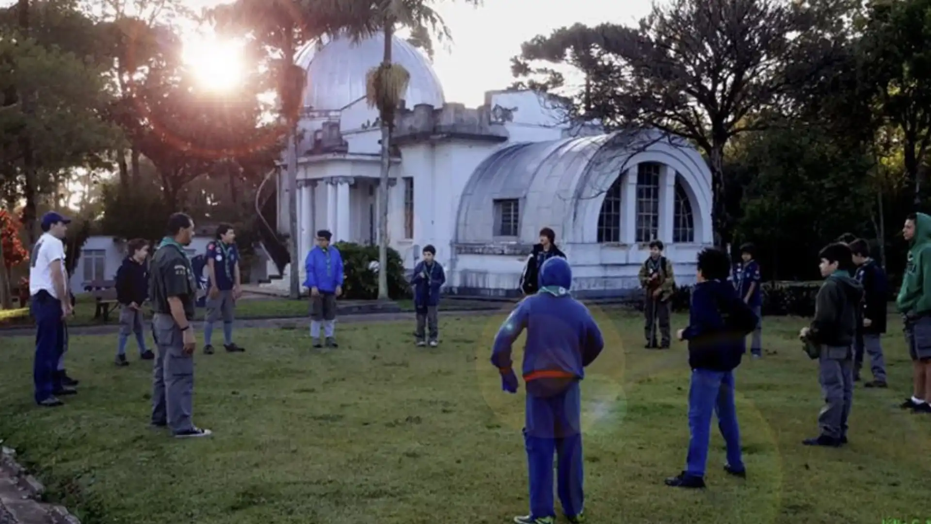 Grupo de jovens escoteiros em um parque com um prédio histórico ao fundo sob a luz do sol poente.