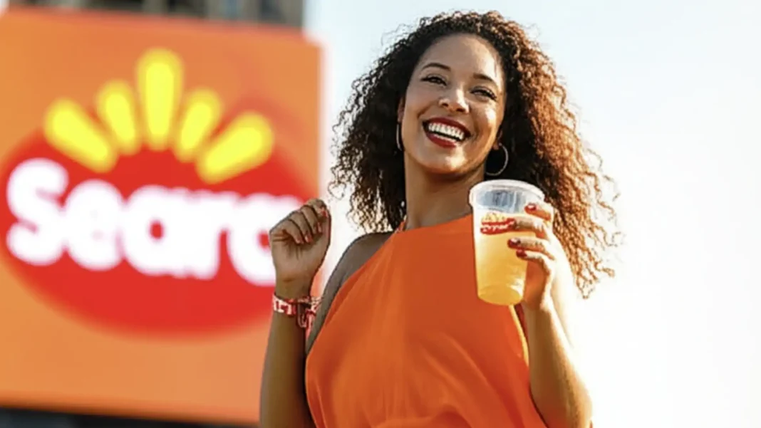 Uma mulher sorridente, com cabelo cacheado e vestindo um top laranja, segura um copo de bebida claro enquanto a marca 