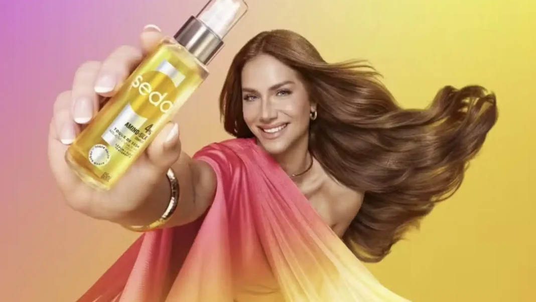 Uma mulher sorridente com cabelos castanhos ondulados segura um frasco dourado de óleo capilar Seda em sua mão esquerda, em frente a um fundo gradiente rosa e amarelo.
