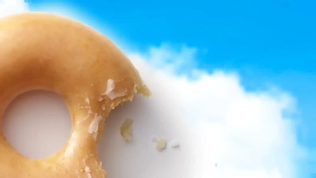 Uma rosquinha da Krispy Kreme com uma mordida, flutuando em frente a um céu azul com nuvens brancas.